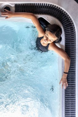 Kabaran bir jakuzide kolları açık dinlenen bir kızın yukarıdan görünüşü. Sıcak jetler ve köpükler uyum, iyileşme ve günlük bakım sağlar..