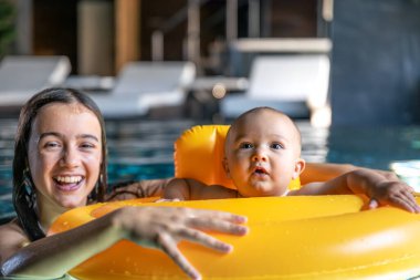 Bir gencin ve parlak sarı şamandıralı bir bebeğin yakın görüntüsü. Köpüklü su ve kahkahalar güvenli oyun, özen ve sıcak bir aile havası sergiliyor..