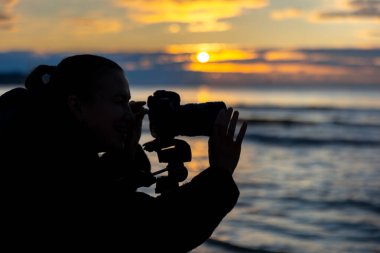 Gölgeli genç bir kadın ufukta güneş batarken bir kamerayla denizi yakalıyor. Sıcak, uyumlu ve yaratıcı bir ortam..