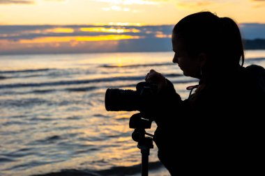 Gün doğumunda deniz kenarında kamerasını tripodla kontrol eden genç bir kadının silueti. Yaratıcılık, içerik yaratma ve gençlik yaşam tarzı..