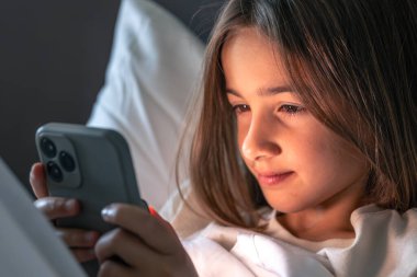 Beyaz çarşaflı gülümseyen bir kızın düşük ışıkta telefonunu okuması. Neşeli ruh hali, olumlu duygular ve rahat bir içtenlik..