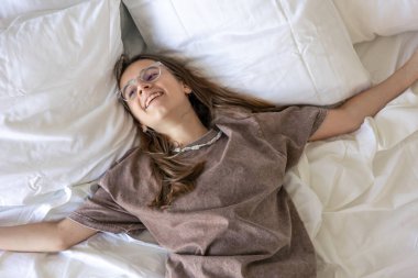 Gözlüklü genç, rahatlayarak gülümseyen yumuşak beyaz yastıkların üzerinde yatıyor. Sıcak ev havası, düşünceli rahatlama ve hafta sonu serinliği..