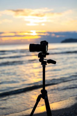 Altın saat boyunca kumlu sahilde bir tripodun üzerindeki kamera, dalgalardaki sıcak yansımalar, sakin bir yaratıcı titreşim oluşturur..