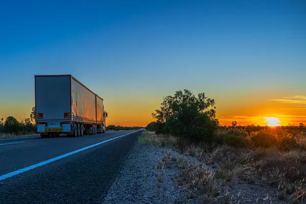 A majestic sunset paints the sky in warm hues of orange and yellow as a semi-truck, labeled as a \'Long Vehicle\', is parked on the side of a quiet highway. The scene captures the tranquil end of a day with the sun dipping below the horizon, casting lo