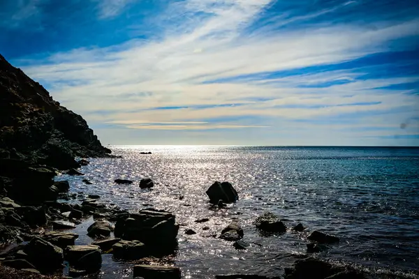 Sunlight dances on the ocean waves, creating a glittering effect against the rocky shore.