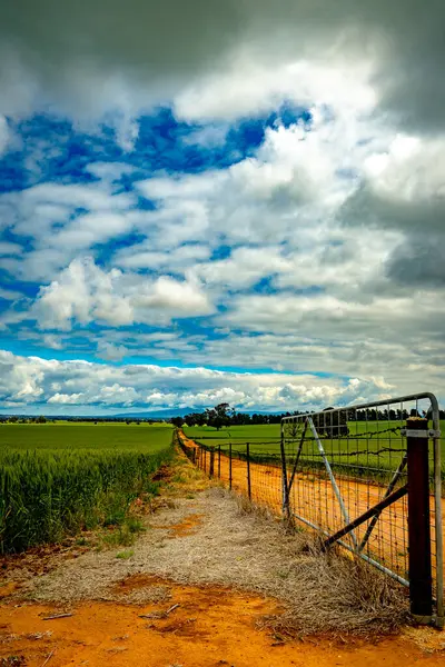 A farm gate opens to a vibrant rural landscape under a sky of sculpted clouds.