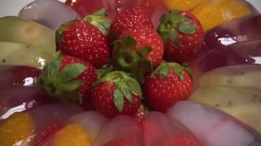 Yuvarlak şekilli sağlıklı meyveli puding beyaz mermer mutfak masasında. Vanilya jölesi, süt, mango suyu ve üzerine taze çilek, üzüm, kivi, mandalina portakalı, hindistan cevizi jölesi (nata de coco) ve taze çilek konmuştur. 4K