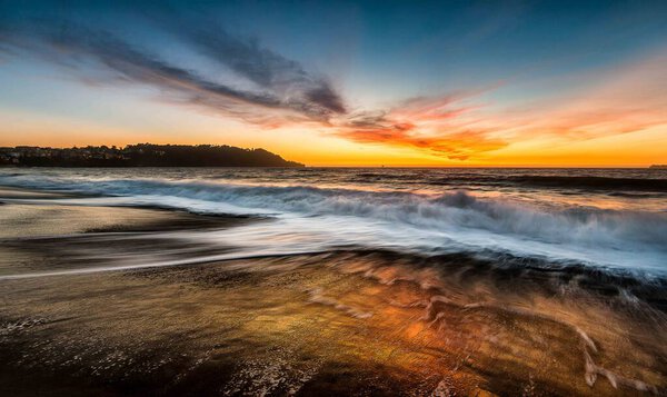 A vibrant, colorful sunset illuminates the ocean as waves crash against the shore.