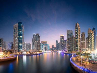Bu görüntü, Dubai Marina 'daki bir şehir silueti. Gökdelenler ve su üzerinde yansıyan binalar yer almaktadır. Aydınlatılmış gökdelenleri ve rıhtımı olan alacakaranlık şehri.
