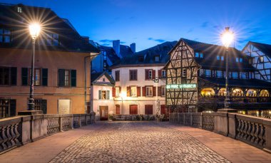 Görüntü, Avrupa 'da gece lambalarıyla aydınlatılmış bir kaldırım taşı caddesini gösteriyor. Geleneksel Alsace mimarisine sahip binalar cadde boyunca uzanır, gölgeler oluşturur.