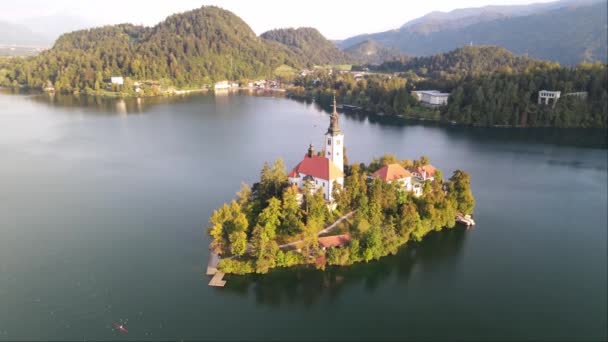 太陽の暖かい輝きを反映した緑と静かな水に囲まれた湖の島に囲まれた歴史的な教会の穏やかな美しさを空想的に示しています — ストック動画