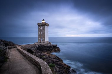 Huzurlu ve pitoresk bir deniz feneri, kasvetli bir gökyüzü ve Brittany Fransa 'da sakin bir deniz tarafından çerçevelenmiş kayalık sahilde görkemli bir şekilde duruyor.