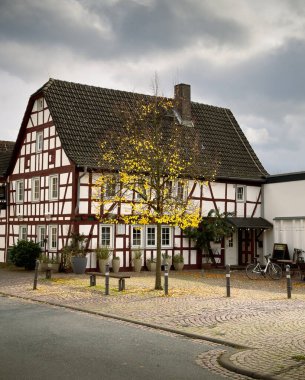 Beyaz duvarları ve kırmızı ahşap çerçeveli eski bir ev kaldırım taşı sokağının önünde duruyor. Evin önünde sarı yapraklı bir ağaç oturuyor..