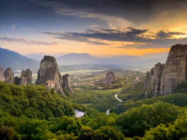 Yunanistan 'ın Meteora bölgesinin uzun ve kayalık bir uçurumun tepesinde tünemiş bir manastır manzarası. Aşağıdaki vadiyi saran yemyeşil arazi boyunca uzanan dolambaçlı bir yol var..