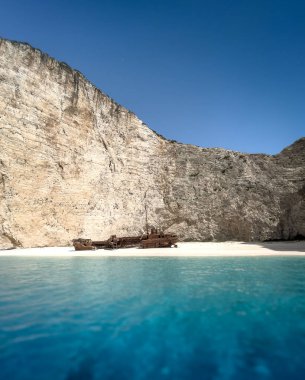 Yunanistan 'ın Navagio Körfezi' ndeki beyaz kumsalda paslı bir gemi enkazı duruyor. Gemi kısmen berrak mavi suya batmış ve arka planda yükselen körfez kayalıkları var..