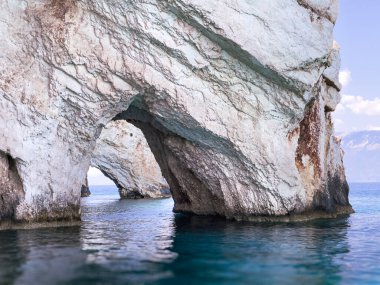 İyon Denizi 'nin üzerinde büyük beyaz bir kaya kemeri duruyor, su berrak bir mavidir. Kemer kıyıya yakın bir yerde ve güneş parlıyor..