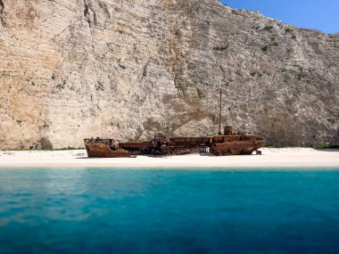 Yunanistan 'ın Navagio Körfezi' ndeki beyaz kumsalda paslı bir gemi enkazı duruyor. Gemi kısmen berrak mavi suya batmış ve arka planda yükselen körfez kayalıkları var..