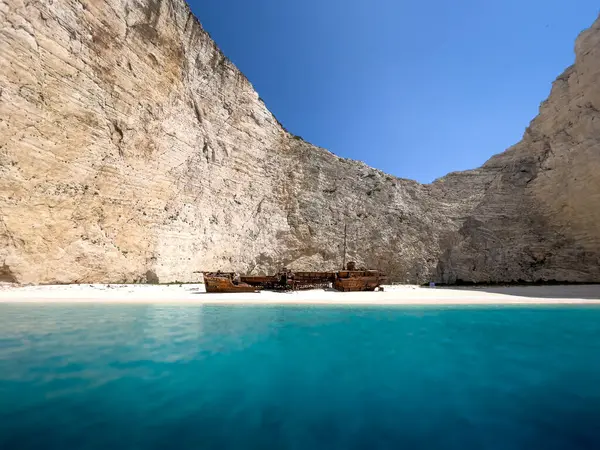 Yunanistan 'ın Navagio Körfezi' ndeki beyaz kumsalda paslı bir gemi enkazı duruyor. Gemi kısmen berrak mavi suya batmış ve arka planda yükselen körfez kayalıkları var..