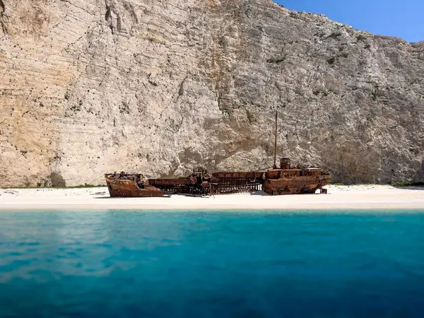 Yunanistan 'ın Navagio Körfezi' ndeki beyaz kumsalda paslı bir gemi enkazı duruyor. Gemi kısmen berrak mavi suya batmış ve arka planda yükselen körfez kayalıkları var..