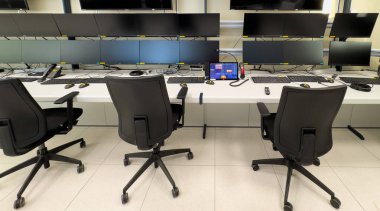 Tekerlekli üç siyah ofis sandalyesi birden fazla bilgisayar monitörlü beyaz bir masaya bakıyor. Monitörlerin hepsi siyah bir ekran gösteriyor. Masada klavye ve fareler var. Ofis aydınlık ve temiz..