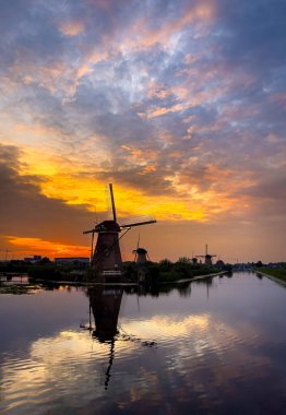 Dramatik bir günbatımı gökyüzüne karşı yel değirmenlerinin manzarası. Yel değirmenleri bir kanalın sakin sularına yansıyor..