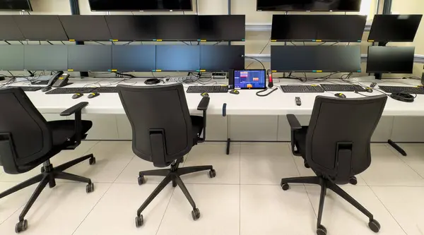 Tekerlekli üç siyah ofis sandalyesi birden fazla bilgisayar monitörlü beyaz bir masaya bakıyor. Monitörlerin hepsi siyah bir ekran gösteriyor. Masada klavye ve fareler var. Ofis aydınlık ve temiz..