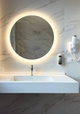 Krom musluğu olan beyaz, dikdörtgen bir lavabo görüntünün merkezindedir. Lavabo beyaz bir tezgahın üzerine monte edilmiştir ve küçük bir dikdörtgen havzası vardır..