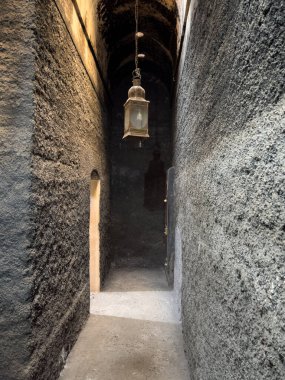 Dar, taş bir koridor tek bir asılı fenerle aydınlatılıyor. Duvarlar sert ve karanlık, zemin de tozlu. Koridorun sonunda bir kapı var ve solda da başka bir kapı var..