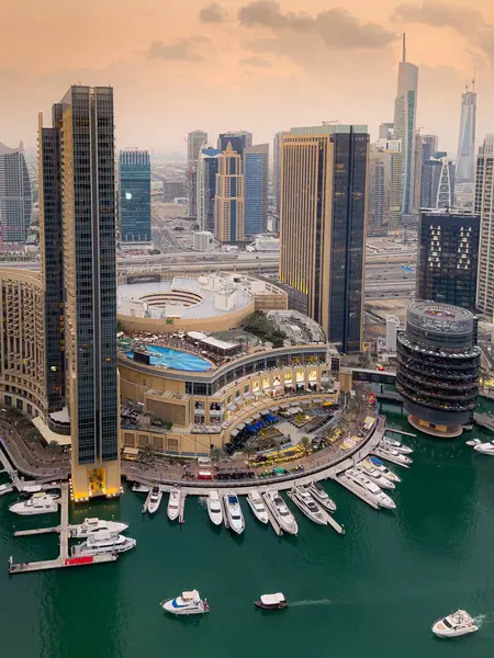 Dubai Marina 'nın kuş bakışı görüntüsü, modern gökdelenler ve lüks dükkanlar sergileniyor. Su durgun, alacakaranlık gökyüzünü yansıtıyor ve kıyı şeridi boyunca bir yat hattı var..