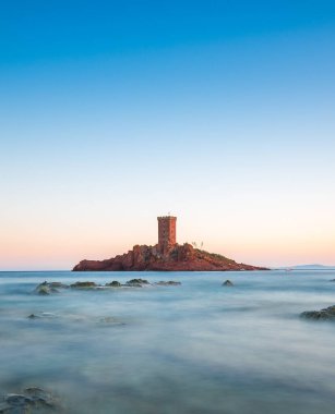 Sakin bir kıyı manzarası, günbatımının ılık parıltısıyla aydınlanan, küçük bir adada yükselen tarihi bir kuleyi, berrak bir gökyüzüne karşı sergiliyor..