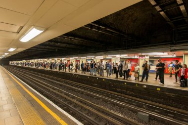 Yoğun bir şehirde kalabalık bir metro istasyonunda bekleyenler var. İnsanlar akşam trafiği sırasında trene binmeye hazırlanırken atmosfer hareketleniyor..