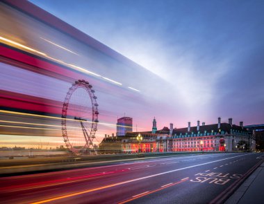 Londra 'da yoğun saatlerde bir otobüsün hareket kabiliyeti bulanıklaşırken, Londra Gözü ve şehir binaları alacakaranlıkta aydınlanıyor. Atmosfer şehrin enerjisini ve enerjisini yakalıyor..