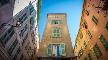 Dar bir caddedeki canlı binaların arasında duran uzun, tarihi bir kule. Parlak yeşil kepenkler pencereleri süslüyor. Güneş ışığı büyüleyici mimariyi aydınlatıyor..