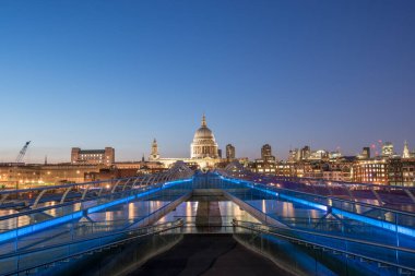 Parlak mavi ışıklar Millennium Köprüsü 'nü süslüyor, St Pauls Katedrali' ne doğru gidiyor ve şehrin ufuk çizgisi alacakaranlıkta parlıyor. Canlı karşıtlık mimari güzelliği vurguluyor.