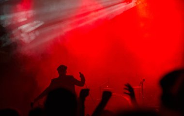 Yoğun kırmızı ışığa karşı silueti olan dinamik bir sanatçı, konser sırasında coşkulu bir kalabalık tezahürat yapıp ellerini kaldırırken,.