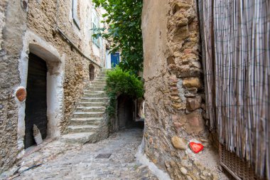 Sessiz bir ara sokakta kaldırım taşları ve antik taş duvarlar, yeşillikle süslenmiş bir merdivene açılıyor. Güneş ışığı ağaçların arasından süzülüp barışçıl bir atmosfer yaratır..