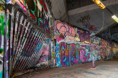 Renkli graffiti, loş ışıklandırılmış bir yeraltı alanının beton duvarlarını süslüyor, çeşitli sanatsal stiller ve mesajlar sergiliyor. Yer, sokak kültürünün kalıntılarıyla dolu..