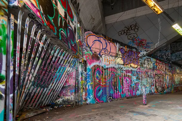 Renkli graffiti, loş ışıklandırılmış bir yeraltı alanının beton duvarlarını süslüyor, çeşitli sanatsal stiller ve mesajlar sergiliyor. Yer, sokak kültürünün kalıntılarıyla dolu..