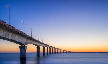 Güzel günbatımı, Ile-de-Re köprüsünün üzerinde ılık renkler bırakır ve sakin sulara doğru zarif bir şekilde uzanır. Yapı, akşam gökyüzüne karşı sokak lambalarıyla aydınlanıyor..
