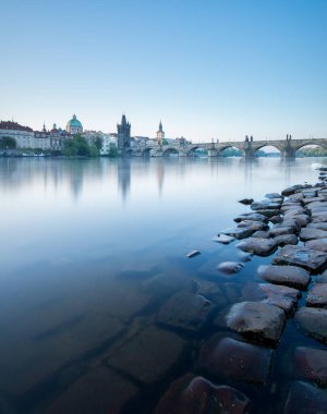 Sabahın erken saatlerinde Vltava Nehri üzerinde sakin bir ortam oluşur, Prag 'ın güzel mimarisini yansıtır ve su boyunca uzanan taştan kıyı şeridini aydınlatır..