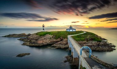 Güneş batarken bir adada duran pitoresk bir deniz feneri, suyun üzerine sıcak renkler döküyor. Karayı deniz fenerine bağlayan uzun bir köprü, dingin atmosferi güçlendirir..