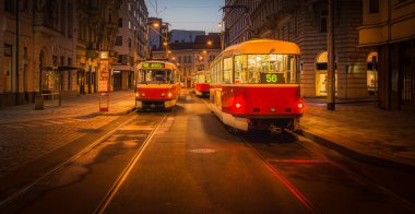 İki tramvay, hareketli bir şehrin sessiz sokaklarında akşam çökerken bir durakta bekliyor. Sıcak ışıklar kaldırım taşlarından yansıyarak sakin bir atmosfer yaratır..