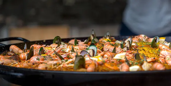 Büyük bir tava paella hareketli bir pazarda karides, midye ve baharat karışımı sergiliyor. Parlak renkler ve cezbedici aromalar bu canlı mutfak ortamında yemek severleri cezbeder..