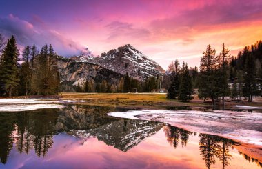 Nefes kesici bir manzara, gün batımında sakin bir dağ manzarasını yakalar. Gökyüzünün canlı renkleri aşağıdaki sakin suda yansıyor, uzun ağaçlarla çevrili..