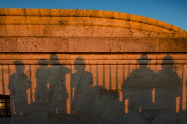 Bir grup insan, güneş batarken siluetleri gerilen taş bir duvarın önünde duruyor. Sıcak ışık duvarın dokusunu aydınlatır ve zengin bir kontrast yaratır..
