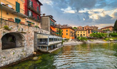 Como Gölü kıyıları boyunca etkileyici binalar dramatik bir gökyüzü altında canlı renkler sergiliyor. Ziyaretçiler göl kenarında geziniyor, sakin atmosferin ve manzaranın tadını çıkarıyorlar..