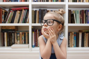 Little cute girl in the glasses sitting in the front of bookshelf. Concept of education in the library