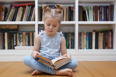 Little cute girl in the glasses sitting in the front of bookshelf. Concept of education in the library