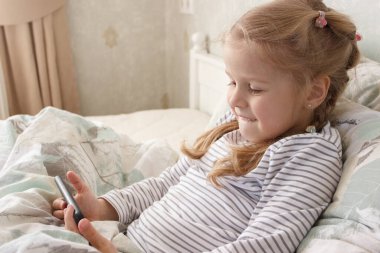 Küçük şirin bir çocuk elinde akıllı bir telefonla yatakta yatıyor. Evde eğitim kavramı