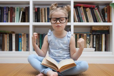 Little cute girl in the glasses sitting in the front of bookshelf. Concept of education in the library